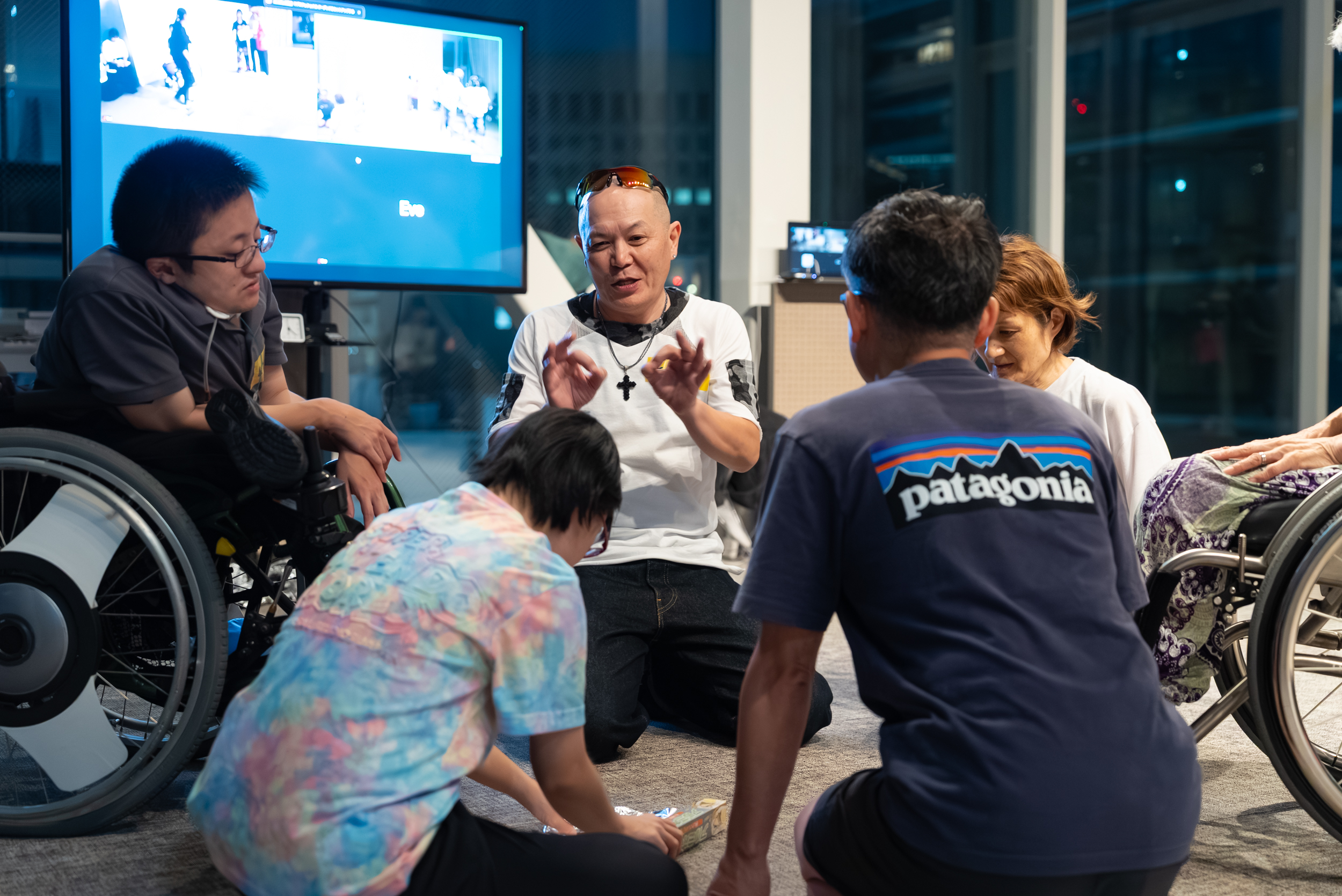 A group of five people, including two wheelchair users, sit together in a circle indoors, engaging in discussion.
