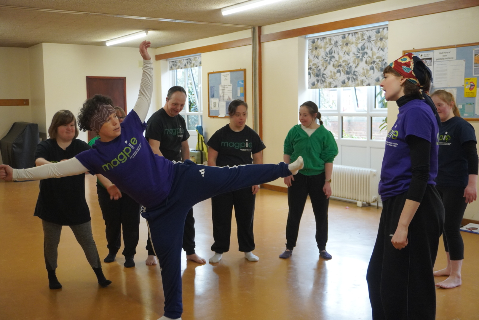 ファシリテーターの一人が片足を高く上げたポーズを取り、周囲の人々が見守りながらダンスの動きを確認している様子。